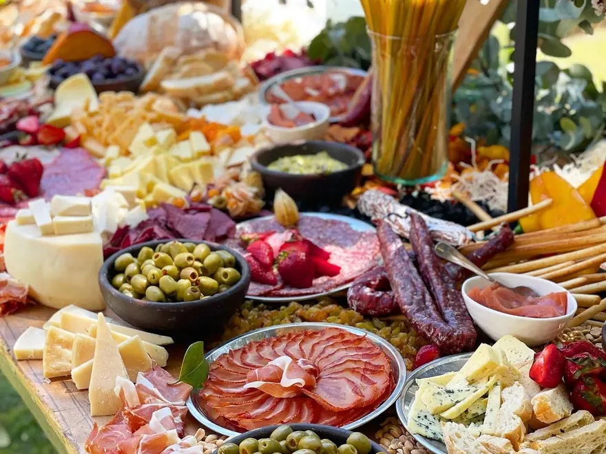 Tabla de charcutería con quesos, embutidos, aceitunas, frutas y grissini montada sobre madera