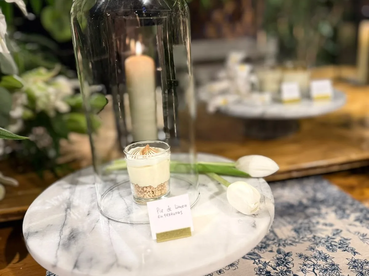 Fanal de vidrio con una vela y un postre de pie de limón rotulado a mano, sobre un pedestal de mármol