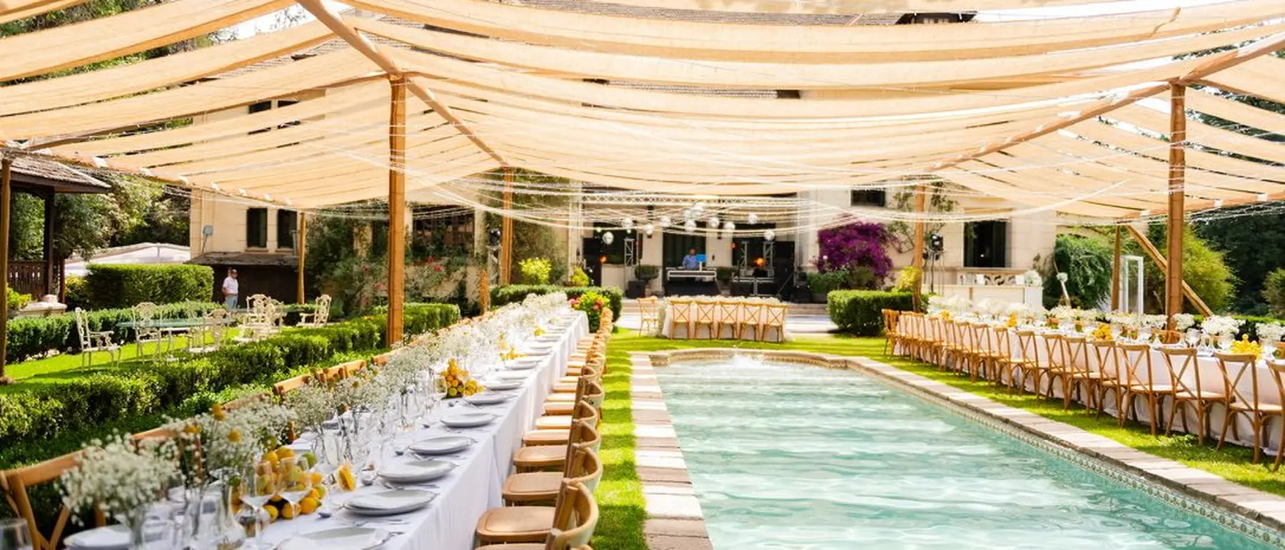 Mesas largas montadas junto a una piscina bajo un toldo, en un matrimonio de día