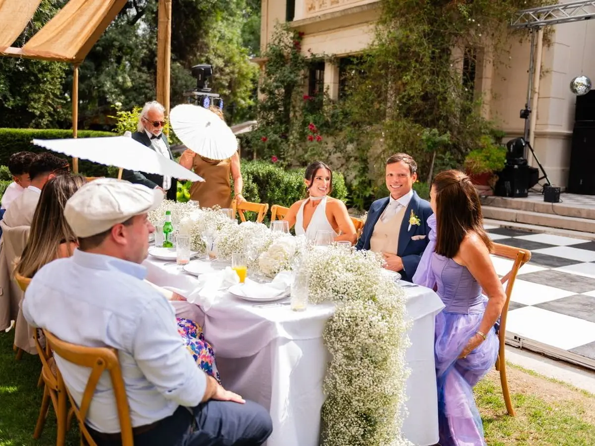 Novios e invitados sentados a una mesa larga con quitasoles, en un matrimonio de día en el jardín