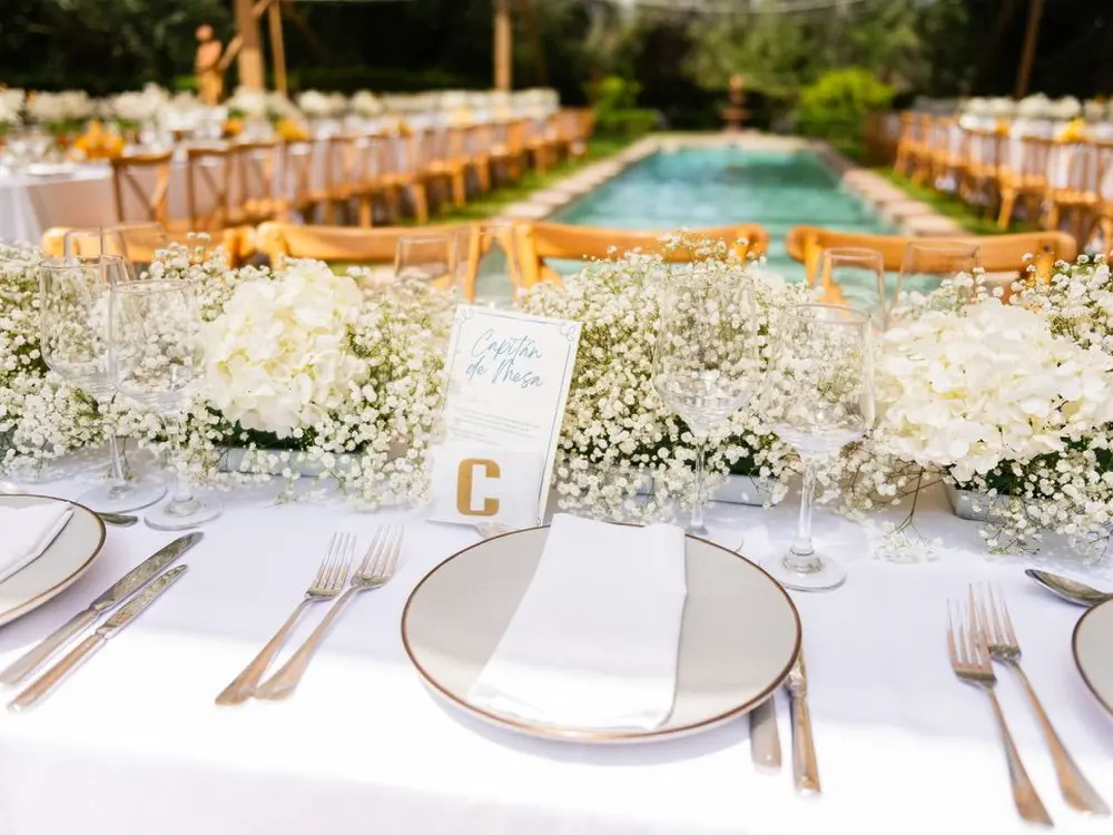 Mesa servida con vajilla, cristalería y flores blancas junto a una piscina