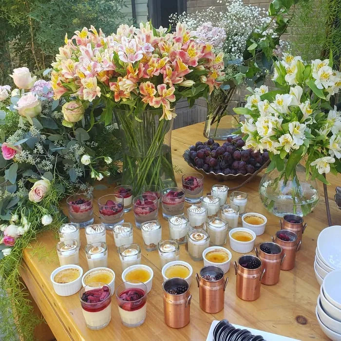 Mesa de postres individuales en vasos, pocillos y jarros de cobre, entre floreros y una fuente de uvas