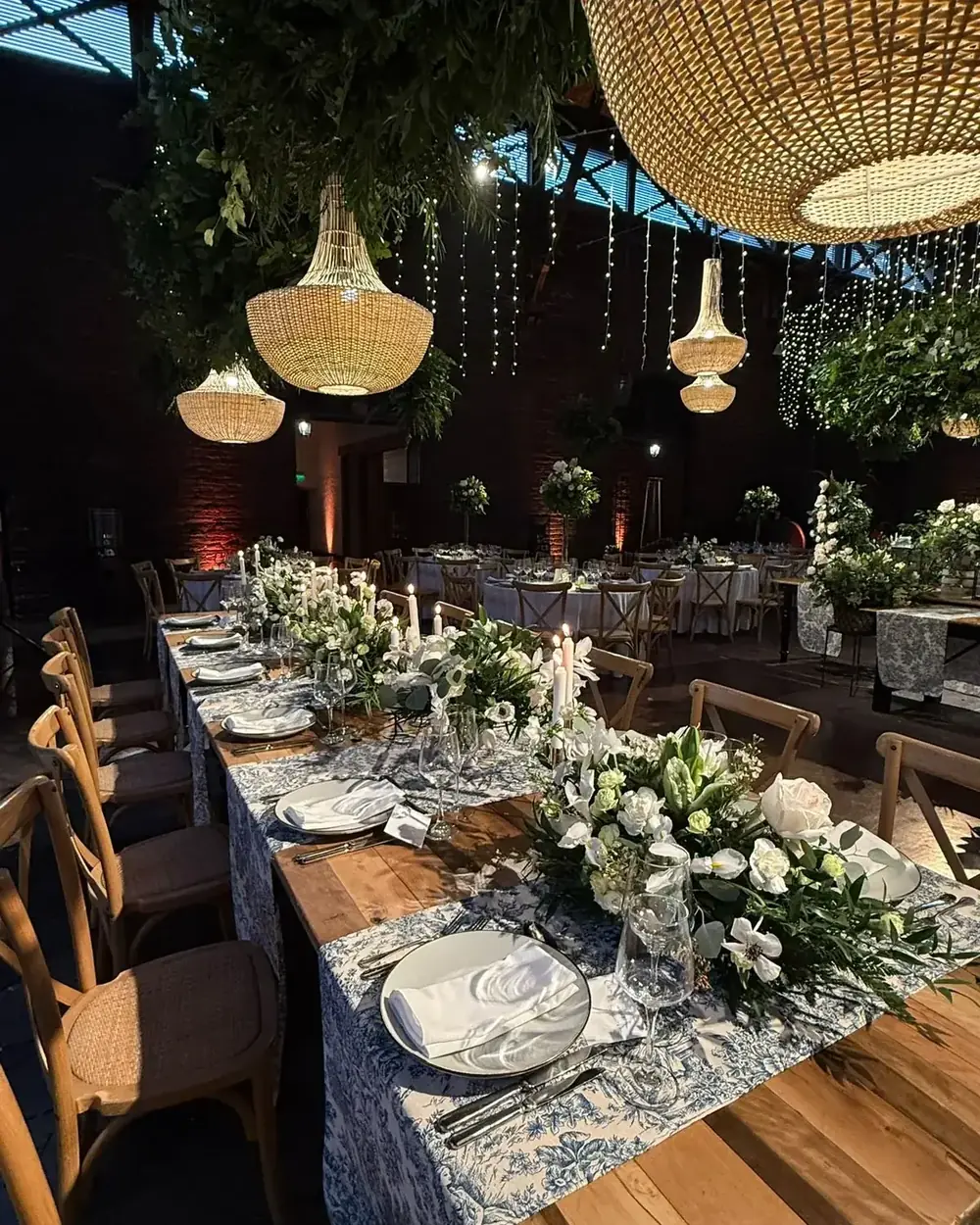 Mesa larga montada bajo lámparas de fibra y follaje colgante, con centros de flores blancas