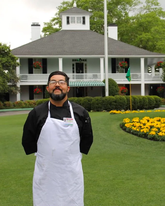 Nicolás Barrios con chaqueta de cocina y delantal frente a la casa club de Augusta National, durante el Masters de 2018