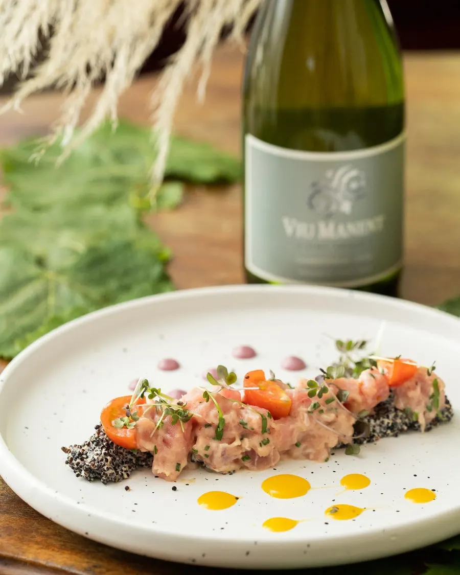 Tártaro emplatado con tomates cherry, semillas y gotas de salsa, junto a una botella de vino